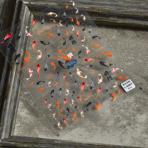 May include: Transparent nail art stickers with koi carp designs in red, orange, black, and white. The sheet includes the words "Lucky Charm" and Chinese characters. A small sticker with the text "to1067" is also visible.