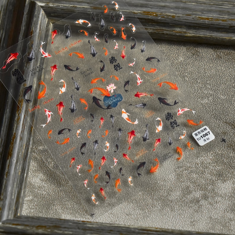 May include: Transparent nail art stickers with koi carp designs in red, orange, black, and white. The sheet includes the words "Lucky Charm" and Chinese characters. A small sticker with the text "to1067" is also visible.
