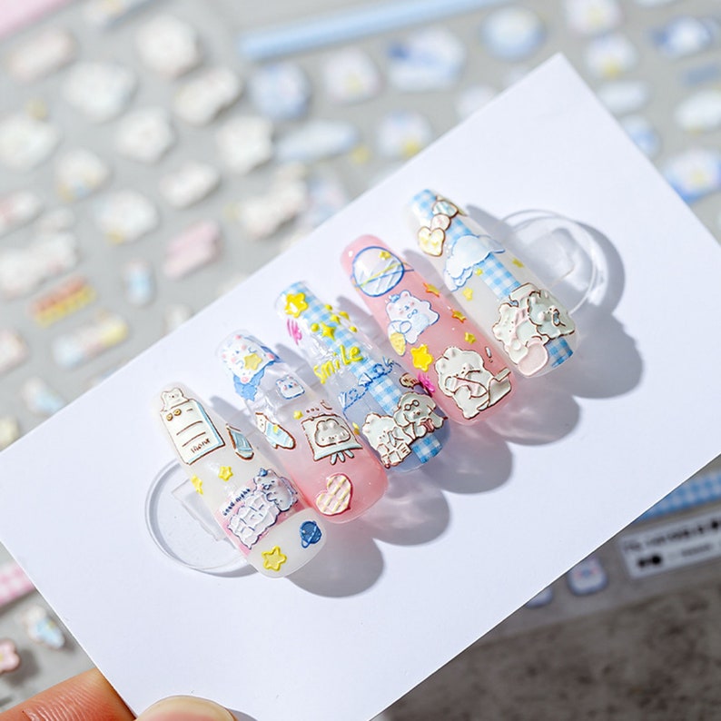 May include: Five artificial nails with cute cartoon designs. The nails feature a variety of pastel colors, including pink, blue, and yellow. Designs include bears, clouds, stars, and hearts. The nails are arranged on a white card.