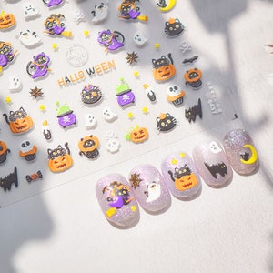 Peut inclure: Une feuille d'autocollants pour ongles d'Halloween avec des chats noirs, des citrouilles, des fant&ocirc;mes et d'autres motifs effrayants. Les autocollants sont sur un fond blanc et disent "Hallo Ween".