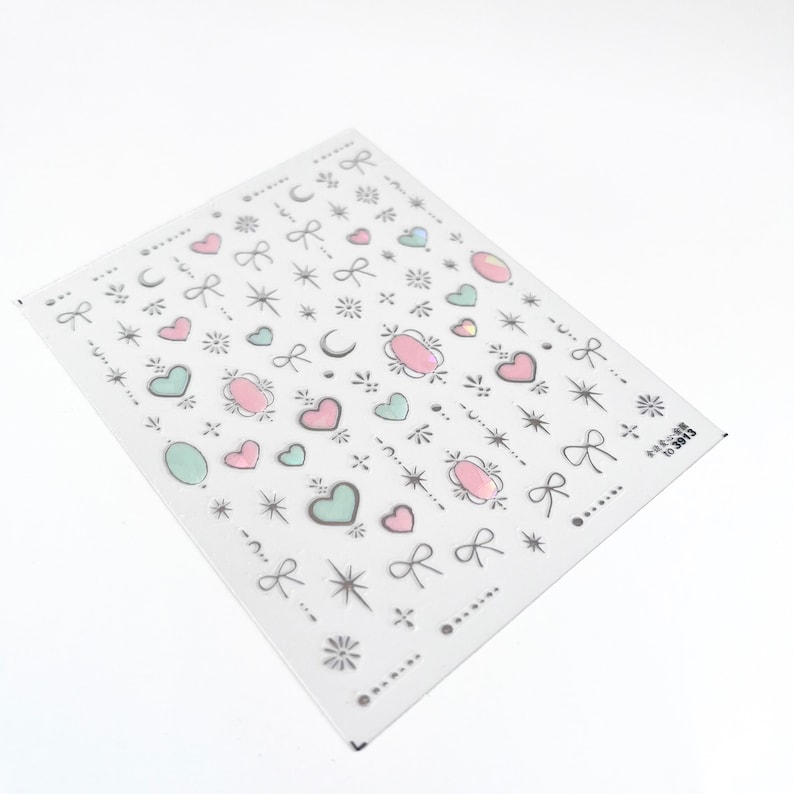 May include: A sheet of nail art stickers with a variety of designs, including hearts, stars, moons, bows, and small oval shapes. The stickers are in shades of pink, green, and silver.