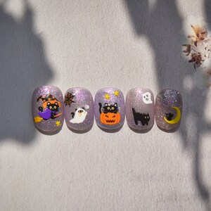 Peut inclure: Cinq designs d'ongles &agrave; paillettes violettes sur le th&egrave;me d'Halloween avec des chats noirs, un fant&ocirc;me, une citrouille, une lune croissante et une toile d'araign&eacute;e.