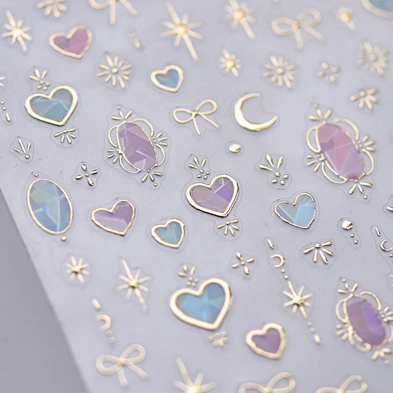 May include: A sheet of iridescent stickers with gold outlines. The stickers feature hearts, stars, moons, bows, and gemstones.