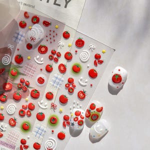 May include: Clear sheet of nail art stickers featuring red tomato designs, smiley faces, bows, and stars. Several manicured nails are decorated with tomato-themed art. The stickers include 3D elements and various tomato sizes.