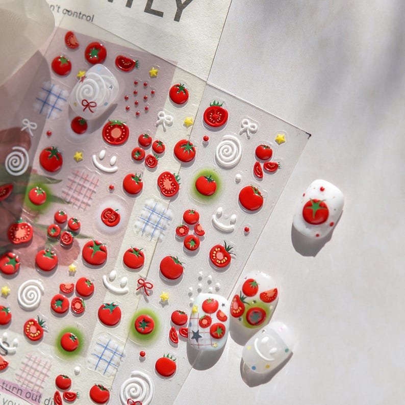 May include: Clear sheet of nail art stickers featuring red tomato designs, smiley faces, bows, and stars. Several manicured nails are decorated with tomato-themed art. The stickers include 3D elements and various tomato sizes.