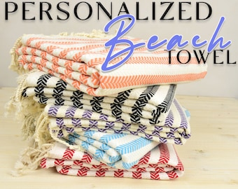May include: A stack of six personalized beach towels in various colors and patterns, including orange, black, purple, blue, and red. The towels are folded and stacked on a light wood surface. The text "Personalized Beach Towel" is visible at the top of the image.