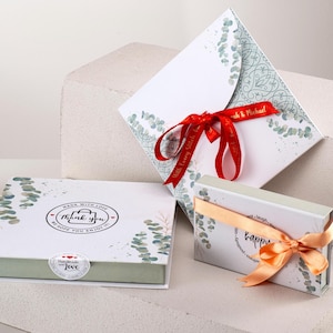 May include: Two gift boxes with a eucalyptus leaf design. The larger box has a red ribbon and the text "Made with Love, Thank You, Hope you enjoy" on the lid. The smaller box has an orange ribbon and the text "Happy" on the lid.