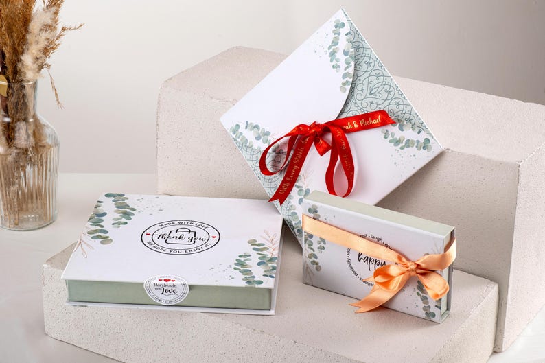 May include: Two gift boxes with a eucalyptus leaf design. The larger box has a red ribbon and the text "Made with Love, Thank You, Hope you enjoy" on the lid. The smaller box has an orange ribbon and the text "Happy" on the lid.