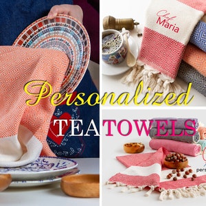 May include: A collection of personalized tea towels in various colors, including coral, red, and blue. The towels feature a geometric diamond pattern and are displayed with kitchenware and food items. Some towels are personalized with the name "Maria".