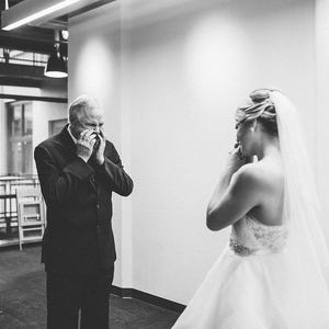 May include: A man in a suit is wiping away tears as he looks at a woman in a white wedding dress. The woman is standing with her back to the camera, and she is holding a bouquet of flowers.