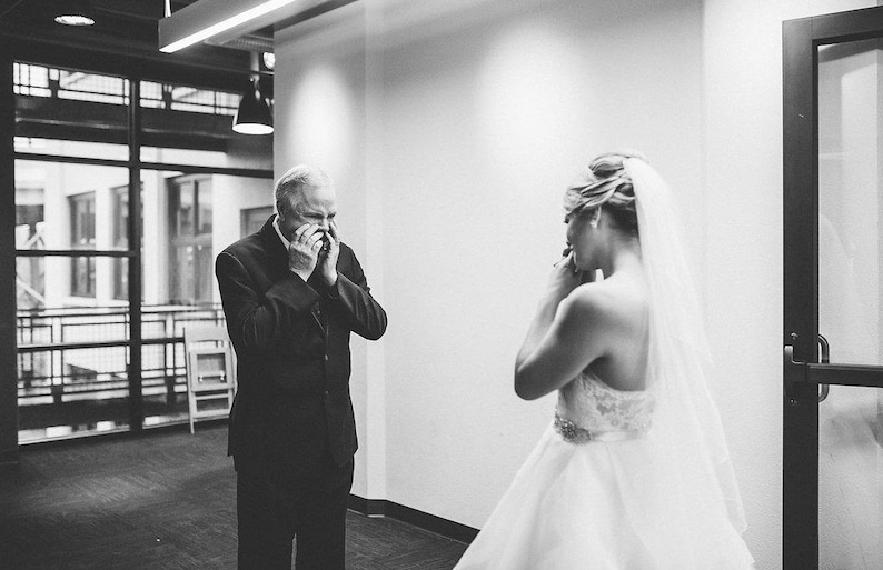 May include: A man in a suit is wiping away tears as he looks at a woman in a white wedding dress. The woman is standing with her back to the camera, and she is holding a bouquet of flowers.