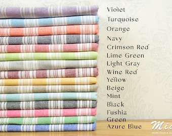 May include: A stack of 16 striped towels in various colors, including violet, turquoise, orange, navy, crimson red, lime green, light gray, wine red, yellow, beige, mint, black, fuchsia, green, and azure blue. The towels are folded and stacked on a wooden surface. The text "Mia MORE THAN A GIFT" is visible on the right side of the image.