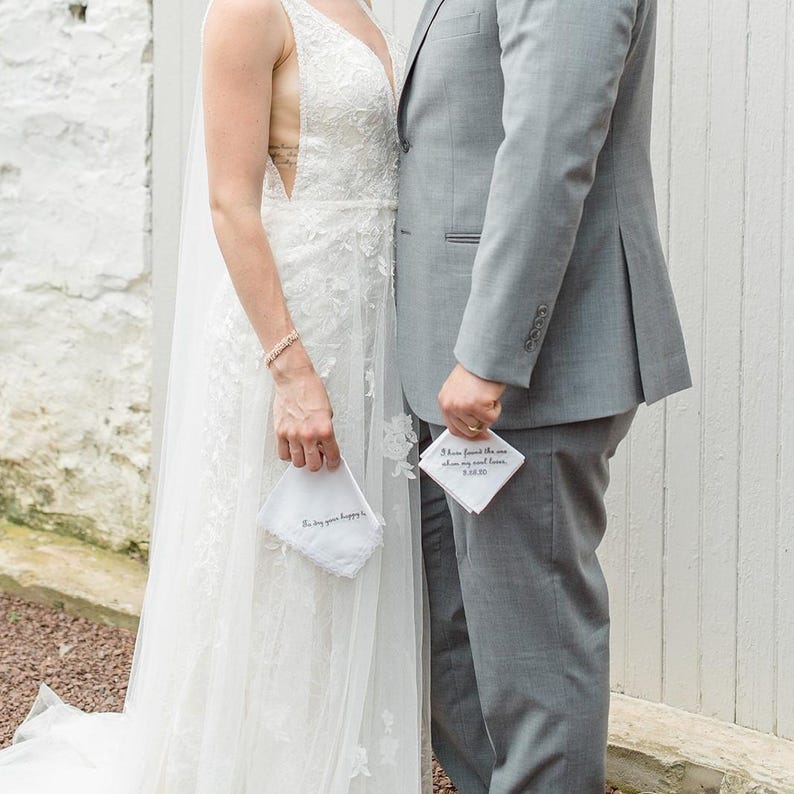 May include: A bride and groom holding white handkerchiefs with personalized messages. The bride is wearing a white lace wedding dress and the groom is wearing a gray suit.