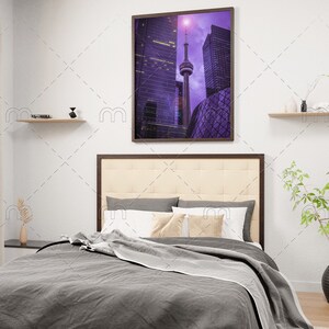 May include: A bedroom interior with a gray bed, white pillows, and a beige headboard. A framed photograph of the Toronto skyline hangs on the wall above the bed. The photograph is in a purple hue. There are two shelves on the wall, one on each side of the photograph. A small plant is on a side table next to the bed.