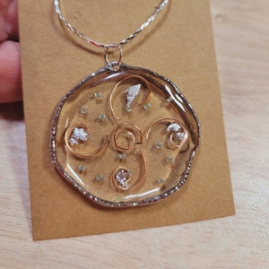 May include: A silver chain necklace with a round, clear resin pendant. The pendant features a gold swirl design with silver and gold glitter accents.