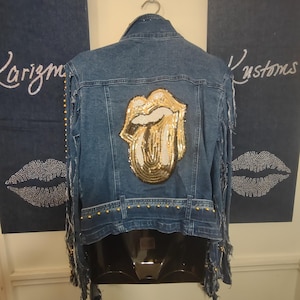 Puede incluir: Chaqueta vaquera azul con un diseño de lengua y labios dorados con lentejuelas en la espalda. La chaqueta tiene tachuelas doradas en la parte inferior y flecos en las mangas. Las palabras "Larizm" y "Kustoms" son visibles en el fondo.