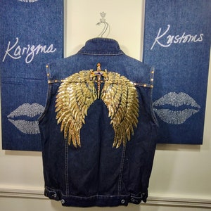 May include: A dark blue denim vest with gold sequined angel wings on the back. The vest has a silver zipper and a silver stud detail. The vest is hanging on a hanger.
