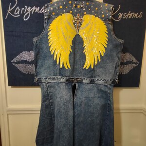 Può includere: Gilet in denim con design di ali d'angelo gialle. Il gilet presenta un lavaggio effetto invecchiato, impreziosito da strass e una croce gioiello. La parte posteriore del gilet presenta grandi ali gialle con paillettes argentate. Il testo "Karizma Kustoms" è visibile.