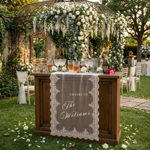 May include: A wooden bar with a lace runner reading "Cheers to The Williams" is set up outdoors. The bar is decorated with champagne, glasses, and floral arrangements. The background features a floral arch and greenery, creating an elegant setting.