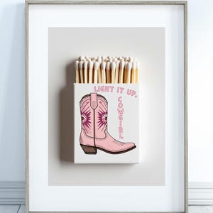 May include: A framed print featuring a matchbox with matches and a pink cowboy boot illustration. The text "LIGHT IT UP, COWGIRL" is displayed on the matchbox. The boot has a floral design.