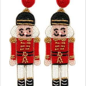 May include: A pair of red, white, black, and gold beaded nutcracker earrings. The nutcrackers are wearing red jackets with gold trim and black hats.