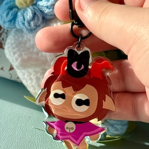 May include: A clear acrylic keychain featuring a cartoon character with red horns, brown hair, and a pink outfit. The keychain has a black clasp and a black bow on the character's head. The character has a stern expression.