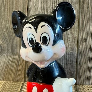 以下が含まれることがあります： ミッキーマウスのセラミック製フィギュア。彼は右手を伸ばして立っており、トレードマークの赤いショートパンツに白い水玉模様を付けています。黒い靴は茶色です。