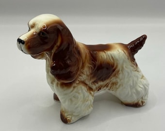 May include: A ceramic figurine of a brown and white cocker spaniel dog. The dog is standing with its head turned to the left and its tail curled up.