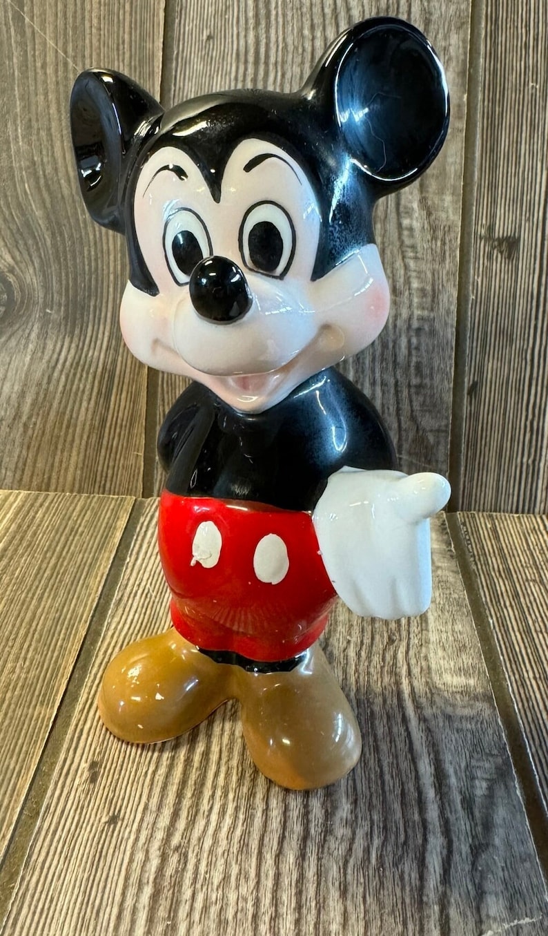 以下が含まれることがあります： ミッキーマウスのセラミック製フィギュア。彼は右手を伸ばして立っており、トレードマークの赤いショートパンツに白い水玉模様を付けています。黒い靴は茶色です。