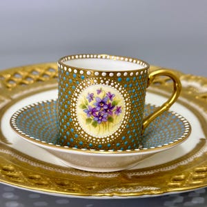 Può includere: Un set da tè in porcellana bianca con bordo dorato e motivo a pois blu e dorati. La tazza presenta un disegno floreale dipinto a mano con violette viola.