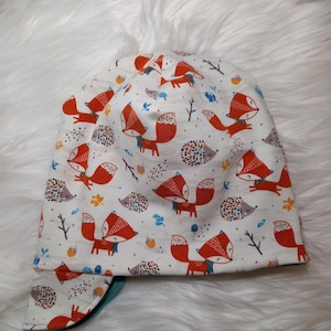Könnte beinhalten: Eine weiße Mütze mit einem Fuchs- und Igelmuster. Die Mütze hat einen umklappbaren Rand in Tealblau.