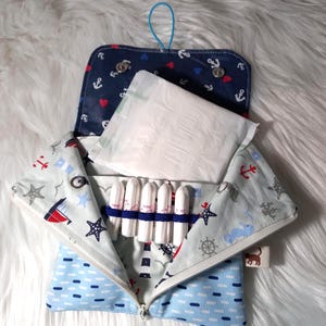 Könnte beinhalten: Geöffnete nautisch thematisierte Tasche mit blauer Schlaufe, mit einem marineblauen Deckel mit roten Herzen und Ankern. Im Inneren sind eine weiße Binde und Tampons zu sehen. Die Tasche hat einen Reißverschluss und ein hellblaues und weißes Muster.