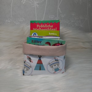 Könnte beinhalten: Ein weißer Stoffaufbewahrungskorb mit rosa Oberseite und einem bunten Design mit Bären und Tipis. Der Korb ist mit einem Stapel Kinderbücher gefüllt, darunter eines mit dem Titel "Fröhliche Weihnachten!"