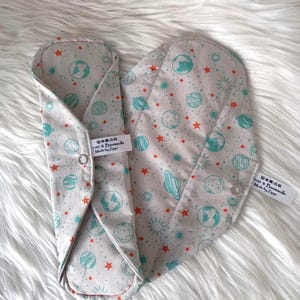 Könnte beinhalten: Zwei wiederverwendbare Stoffbinden mit hellgrauem Hintergrund. Die Binden sind mit türkisfarbenen Planeten, orangefarbenen Sternen und Sonnen gemustert. Jede Binde hat einen Druckknopfverschluss. Hergestellt aus 100 % Baumwolle.