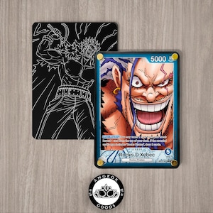 May include: A collectible trading card featuring Rocks D. Xebec with a power level of 5000, displayed in a protective case with gold corner screws. Beside it is a black card back with a white line art illustration of a muscular character.