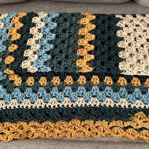 May include: A folded, handmade crochet blanket in a granny square pattern. The blanket features a color palette of teal, light blue, tan, and mustard yellow. The edges are scalloped, and the blanket is folded on a gray surface.