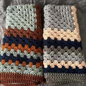 May include: Two folded crochet blankets featuring a granny square pattern. The left blanket displays stripes of sage green, dark brown, navy blue, and burnt orange. The right blanket features stripes of light gray, cream, navy blue, and dark gray.