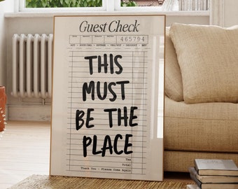 May include: A black and white printable wall art design with the text "Guest Check" and "This Must Be The Place" on a white background with black lines. The design is framed in a light wood frame.