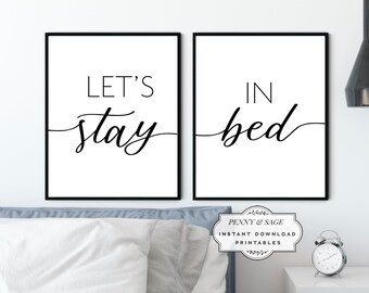 May include: Two black framed prints with white backgrounds. The left print says "Let's stay" in black cursive font. The right print says "in bed" in black cursive font. The prints are on a white wall above a blue bedspread.