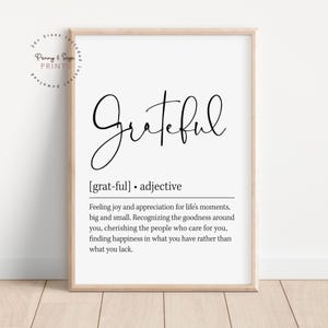 May include: A framed print with the word "Grateful" in elegant black script. Below, the definition of grateful is provided, describing it as an adjective. The print is in a wooden frame, suitable for home decor.