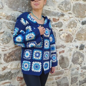May include: A blue crocheted cardigan with a white and blue geometric pattern. The cardigan has a hood and is open in the front. The model is wearing black pants and sunglasses.