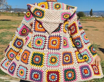 May include: A colorful, hooded crochet jacket featuring a patchwork design of vibrant squares in shades of red, orange, yellow, green, blue, and purple. The jacket has a cream-colored hood and trim, creating a striking contrast against the bright squares. The jacket is worn outdoors.