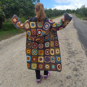 May include: A long, crocheted cardigan with a hood. The cardigan is made of colorful squares with a variety of patterns. The cardigan is worn by a person with long brown hair.