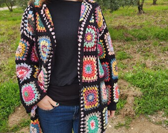 May include: A black crochet cardigan with a colorful granny square pattern. The cardigan has a hood and is open in the front. The person wearing the cardigan is standing in a field.