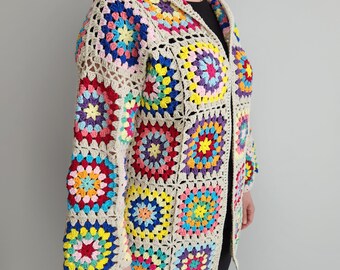 May include: A hooded, open-front crochet jacket featuring a patchwork design. The jacket showcases colorful squares in shades of red, blue, yellow, and purple, set against a neutral beige background. The sleeves and body are constructed from these vibrant, geometric patterns.