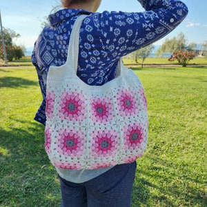 May include: A white crocheted tote bag with pink and gray flower designs. The bag is being worn by a person wearing a blue and white patterned shirt.