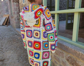 May include: A long, hooded crochet cardigan in a patchwork design. The colorful squares feature various hues, including red, yellow, blue, and green, with a cream-colored hood and trim. The garment is worn outdoors.