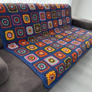 May include: A vibrant, hand-crocheted blanket with a patchwork design. The blanket features squares in shades of blue, orange, yellow, pink, and green. The blanket is draped over a grey sofa, showcasing its intricate pattern and texture.