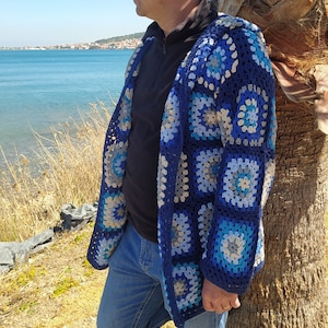 May include: A blue crocheted cardigan with a geometric pattern of squares and circles. The cardigan is made with a combination of blue, white, and gray yarn.