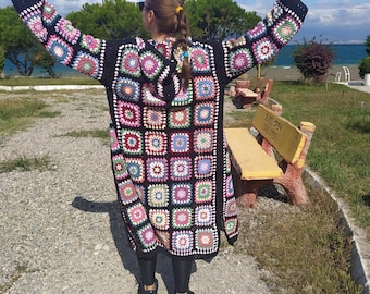 May include: A long, black crochet cardigan with a hood, featuring colorful square patterns. The cardigan has long sleeves and is worn outdoors. The squares are in various colors, including pink, green, and blue.